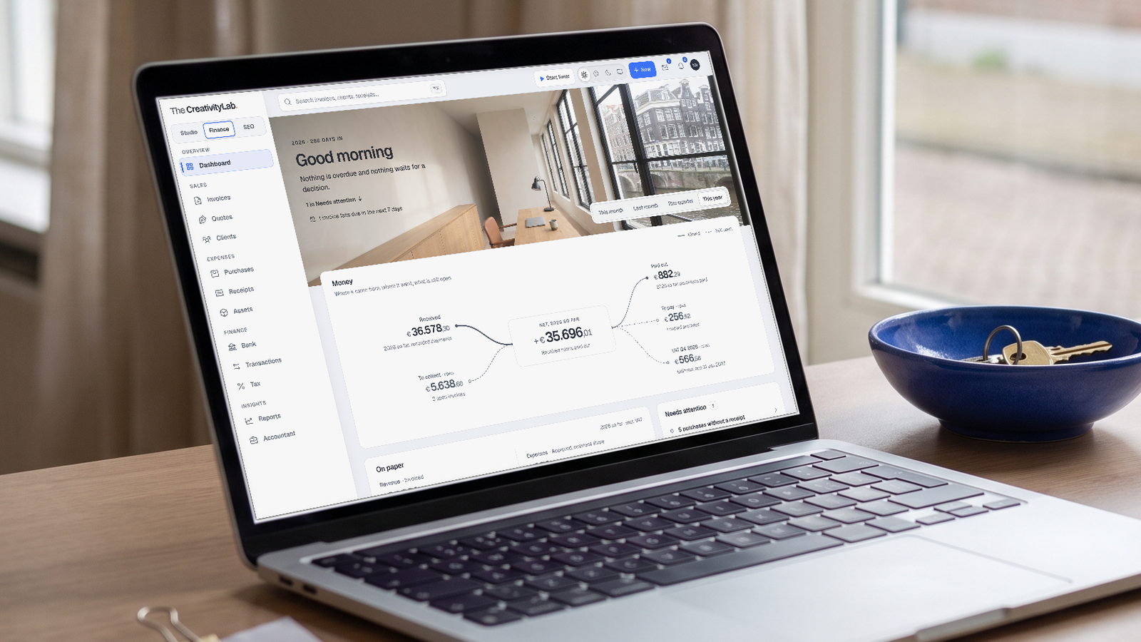 Finance OS: the dashboard of a fictional studio administration on a laptop at a desk by the window, with the year view of money received, open and to pay