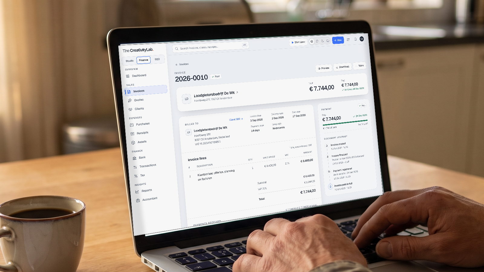 Finance OS: a paid invoice with lines, VAT and payment history on a laptop at the kitchen table, hands on the keyboard