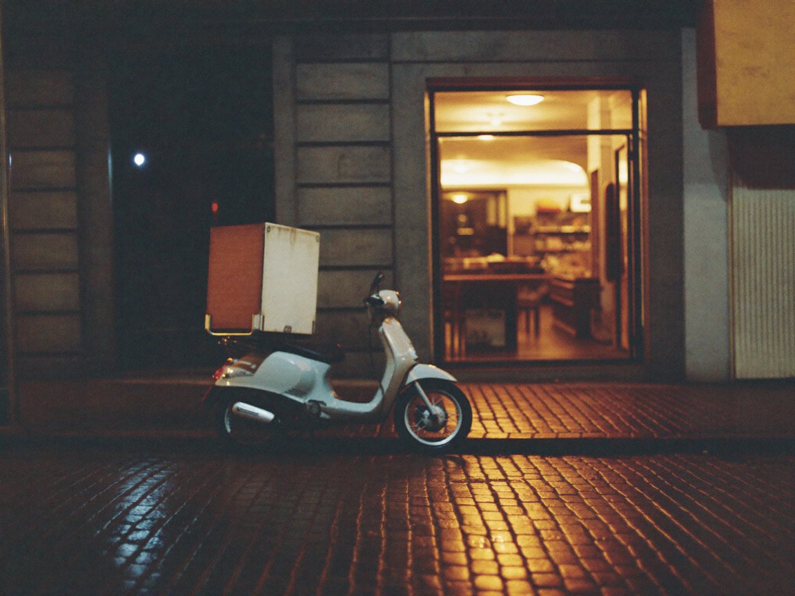 A parked delivery scooter with a delivery box on a wet cobbled street at night.