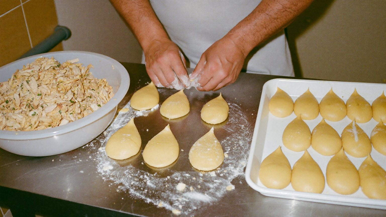 Hands shaping dough into coxinhas on a floured steel worktop, beside a bowl of chicken filling.