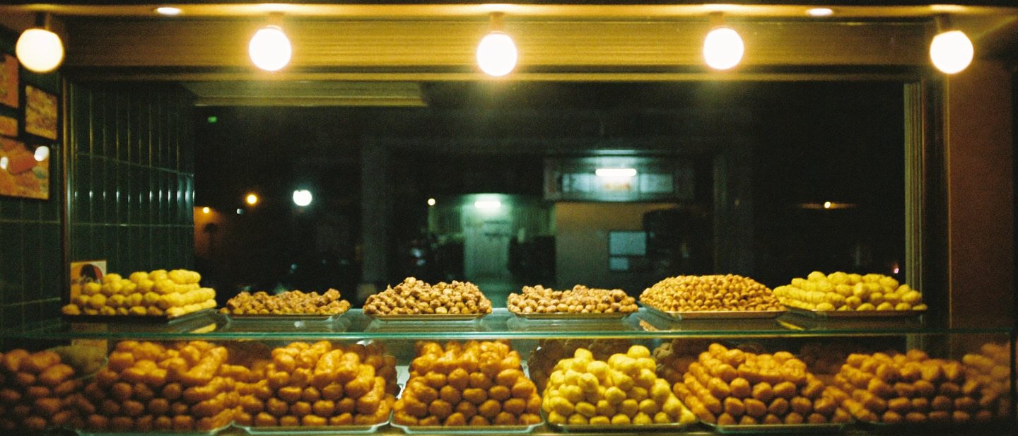 A small snack bar at night with glass trays full of golden coxinhas, warm bulbs and green tiles.