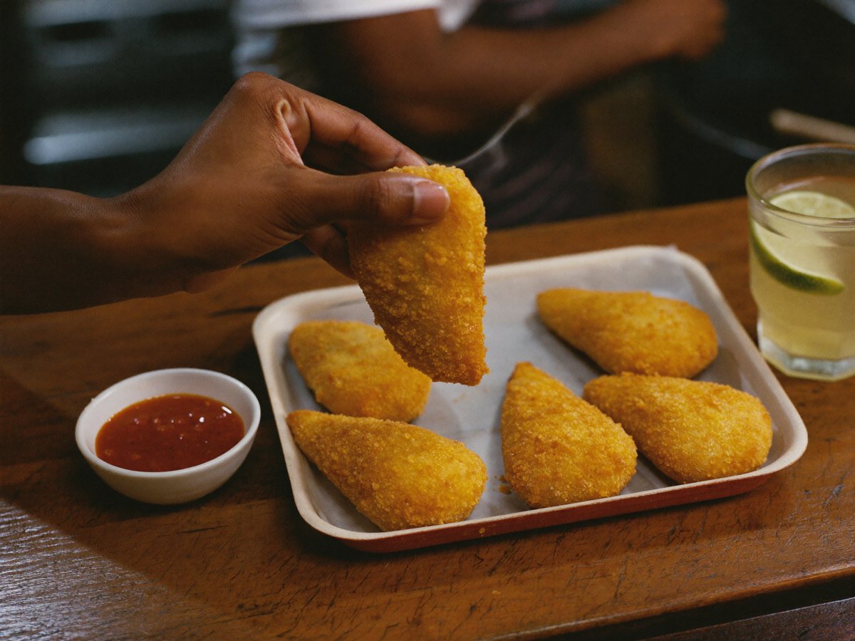 A hand picks up a golden coxinha from a tray with five more, on a wooden counter with a small bowl of chilli sauce and a glass of lime juice in warm evening light.