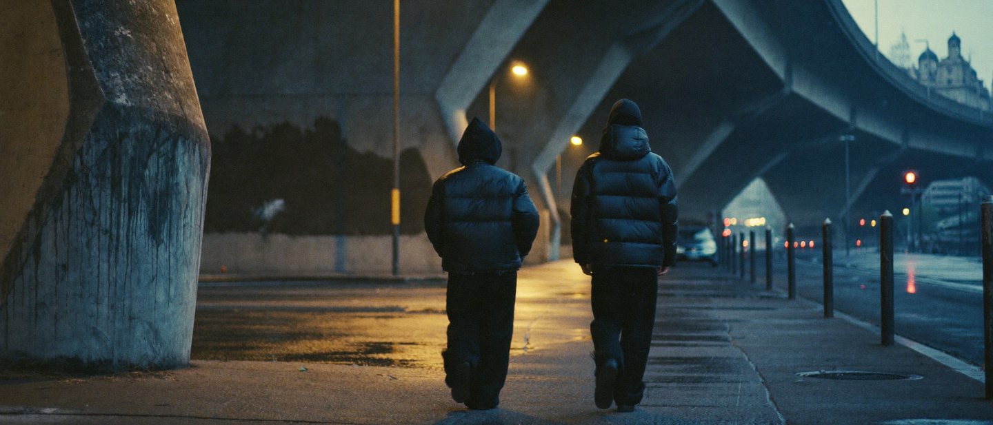 Twee jonge mensen in wijde zwarte streetwear lopen in het blauwe uur over een natte stoep onder een betonnen spoorviaduct.