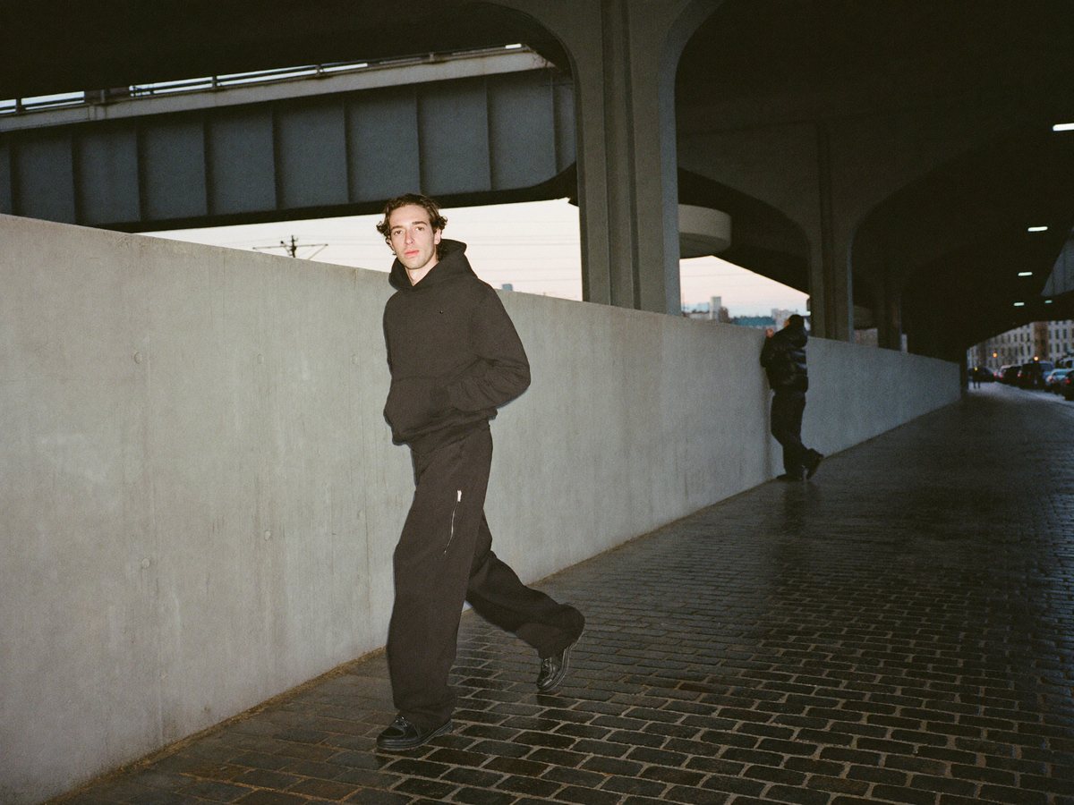 Een jonge man in een oversized zwarte hoodie en wijde zwarte broek loopt in de schemering langs een betonnen muur onder een spoorviaduct, een vriend in een zwarte pufferjas leunt verderop tegen de muur.