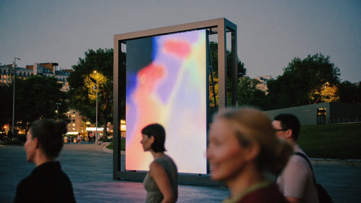Een hoog staand ledscherm met kleurrijk abstract beeld naast een festivalplein in de schemering.