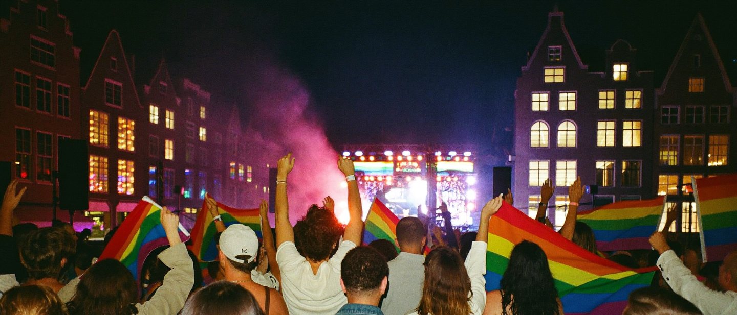 Een grote menigte voor een buitenpodium op een historisch stadsplein in de avond, met gekleurd podiumlicht en regenboogvlaggen.