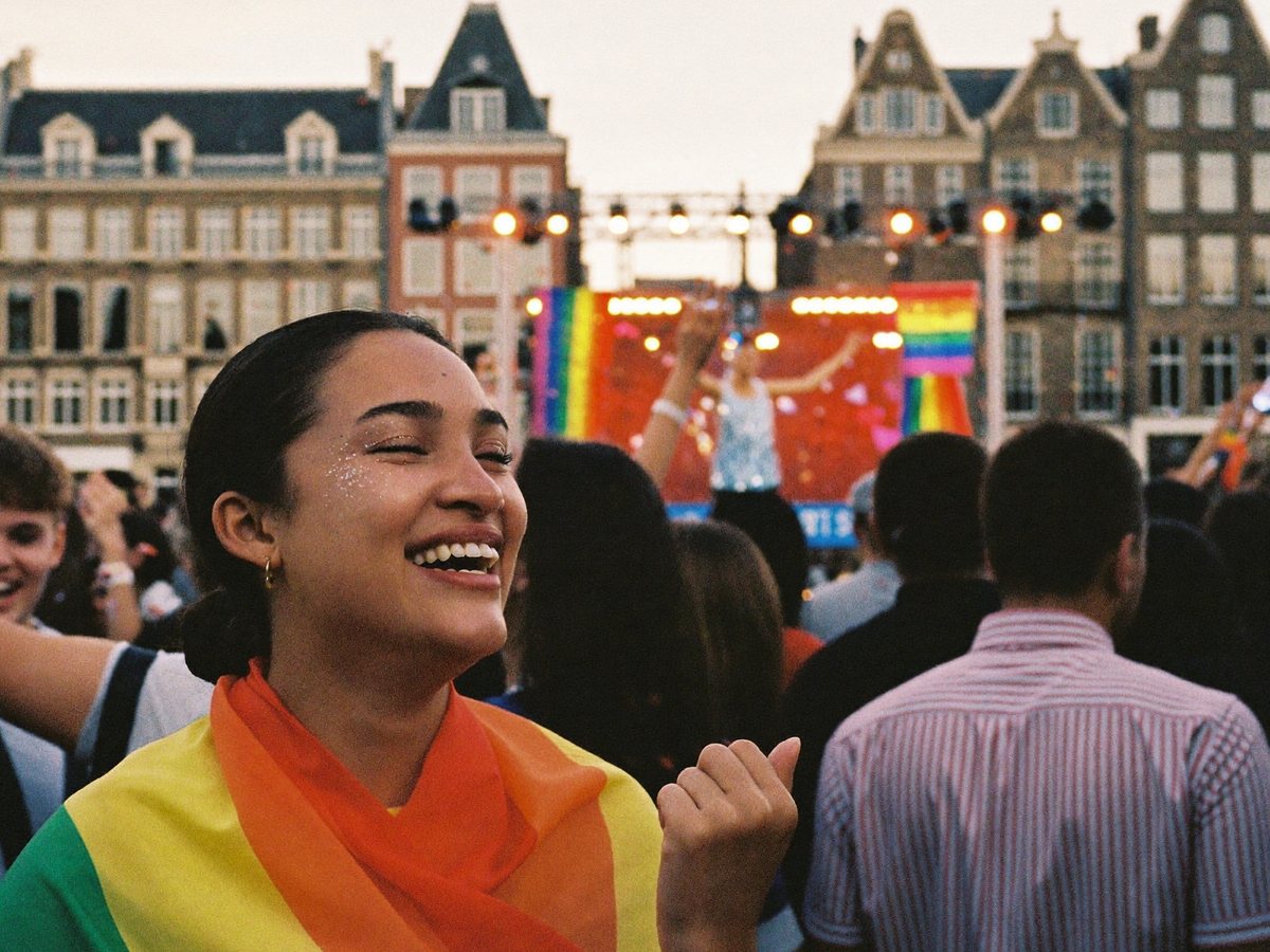 Een lachende jonge vrouw met glitter op haar wangen en een regenboogvlag om haar schouders in de menigte voor een Pride-podium op de Dam, met de Amsterdamse gevels erachter.