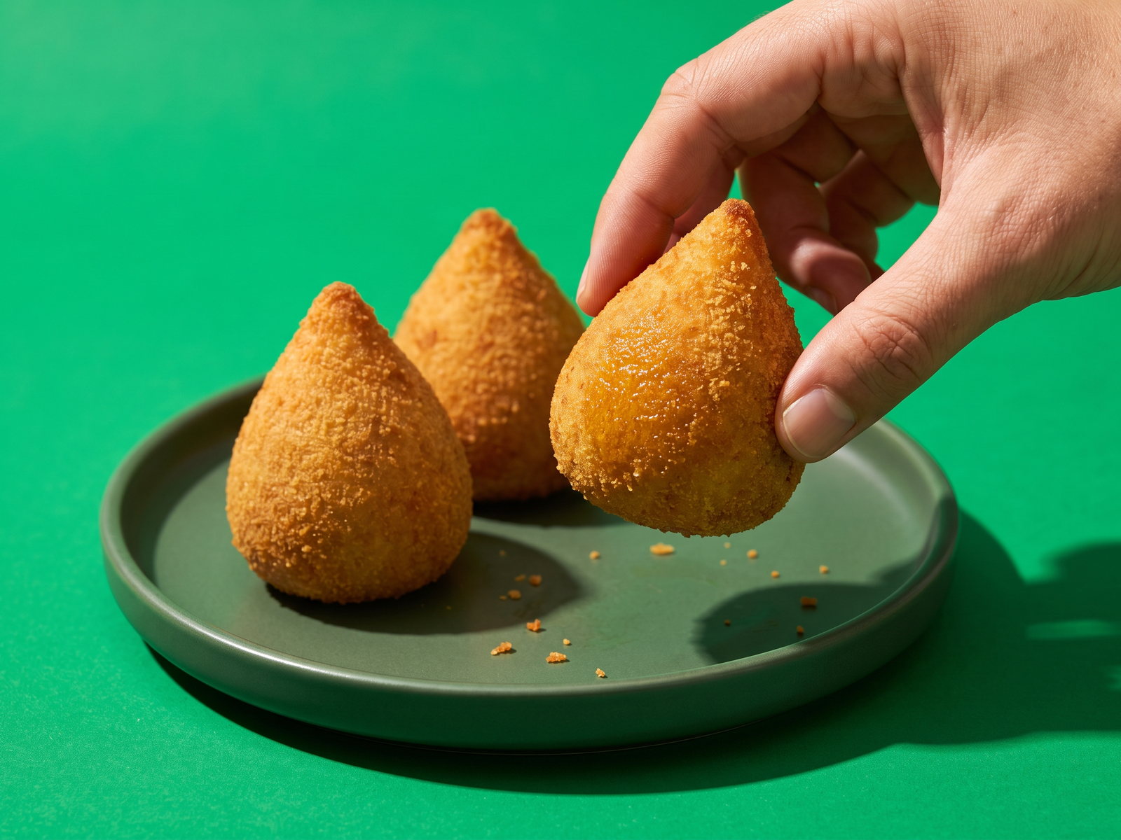 Drie goudbruine coxinha's op een donkergroen bord, een hand pakt er één, tegen een felgroene achtergrond.