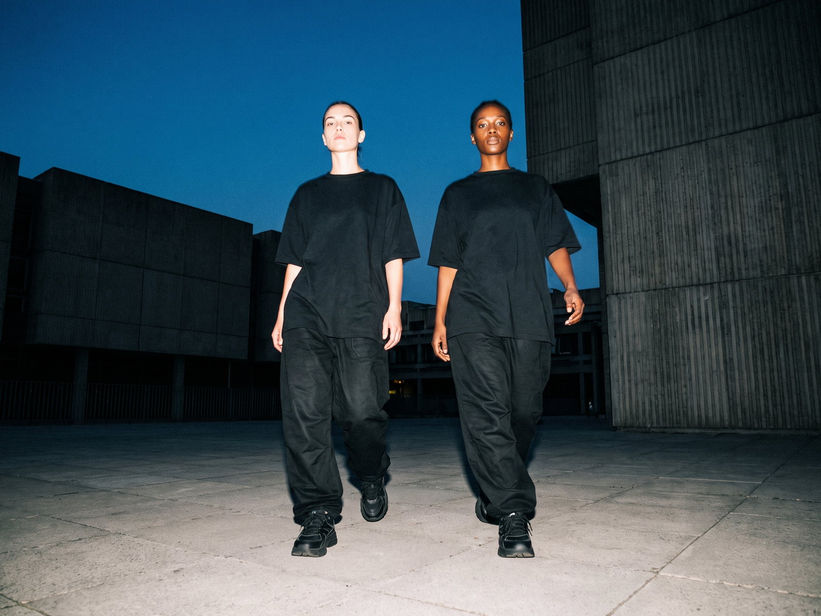 Twee mensen in volledig zwarte oversized outfits lopen tijdens het blauwe uur over een leeg betonnen plein tussen brutalistische gebouwen, bevroren door harde flits.
