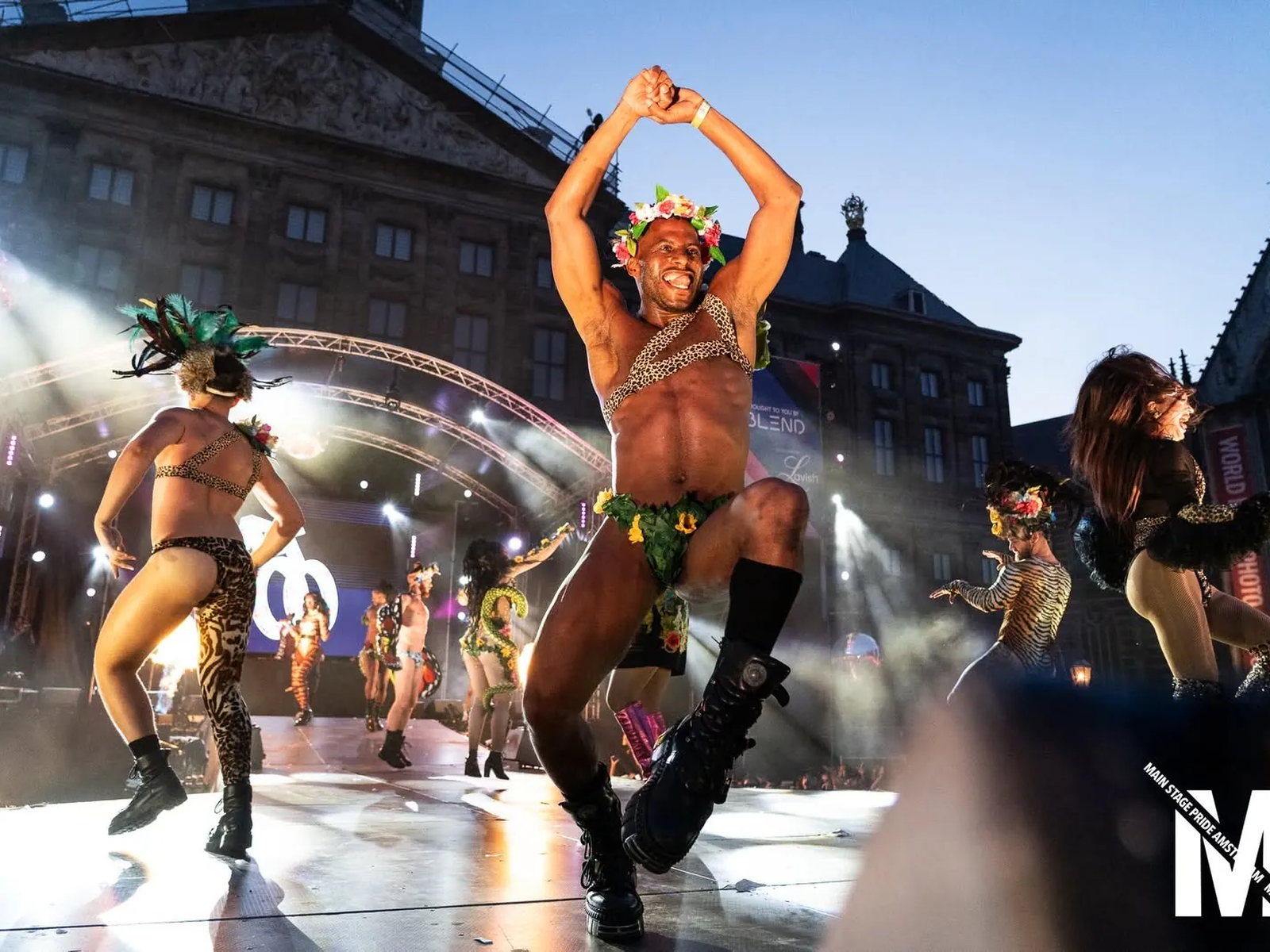 Dansers in luipaardprint en bloemenkransen op het podium van MainStage Pride op de Dam, in het avondlicht voor het Paleis.