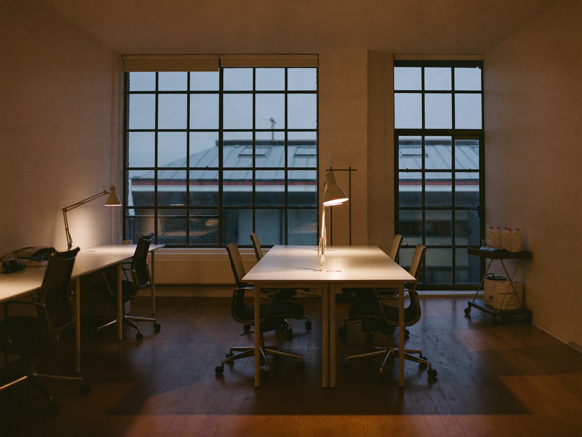 A tidy office in an old warehouse at dusk, with empty desks, lit desk lamps and a cleaning trolley by the window.