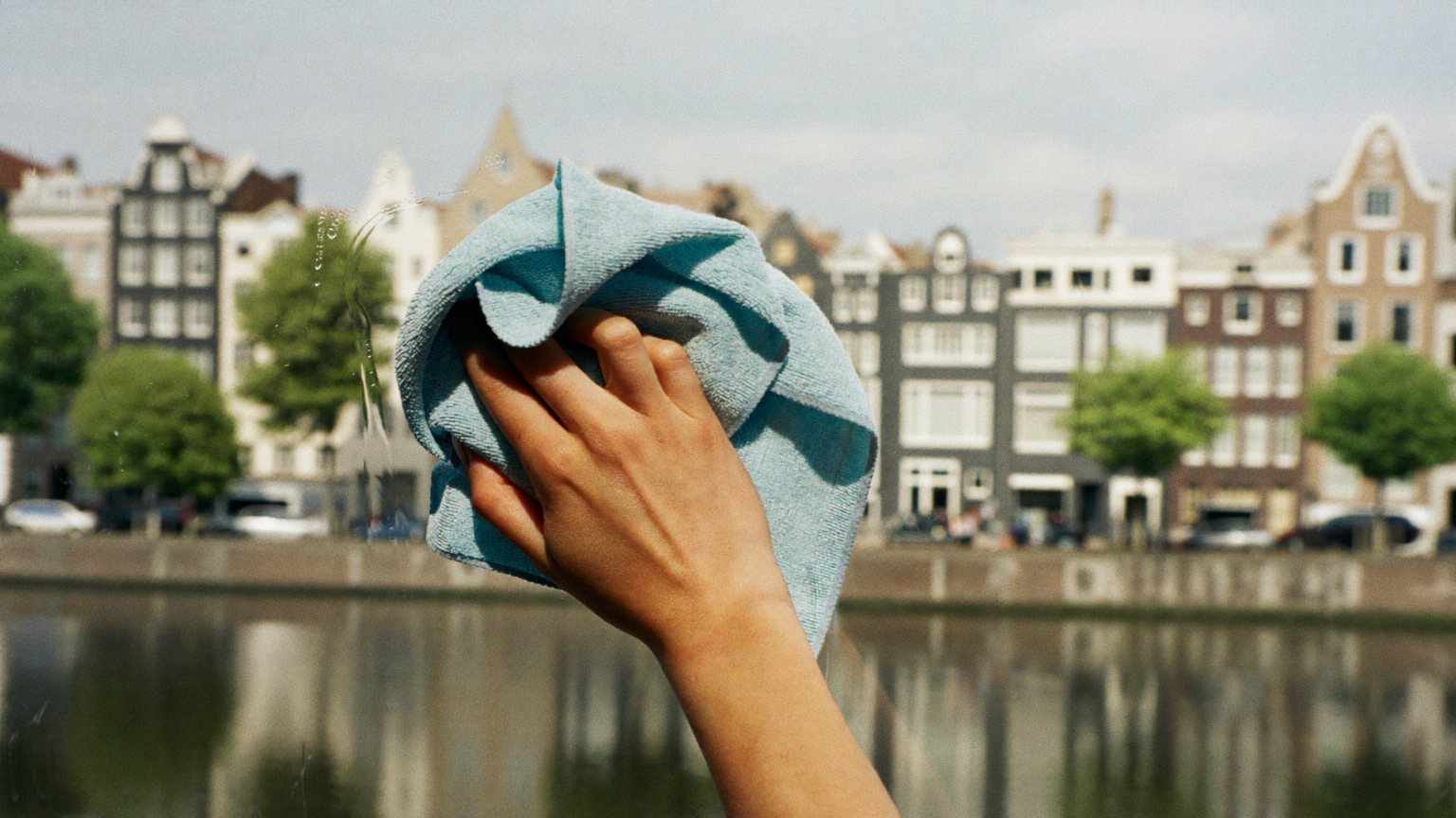 A hand with a light blue microfibre cloth polishes a window, Amsterdam canal houses and their reflection in the water behind it.