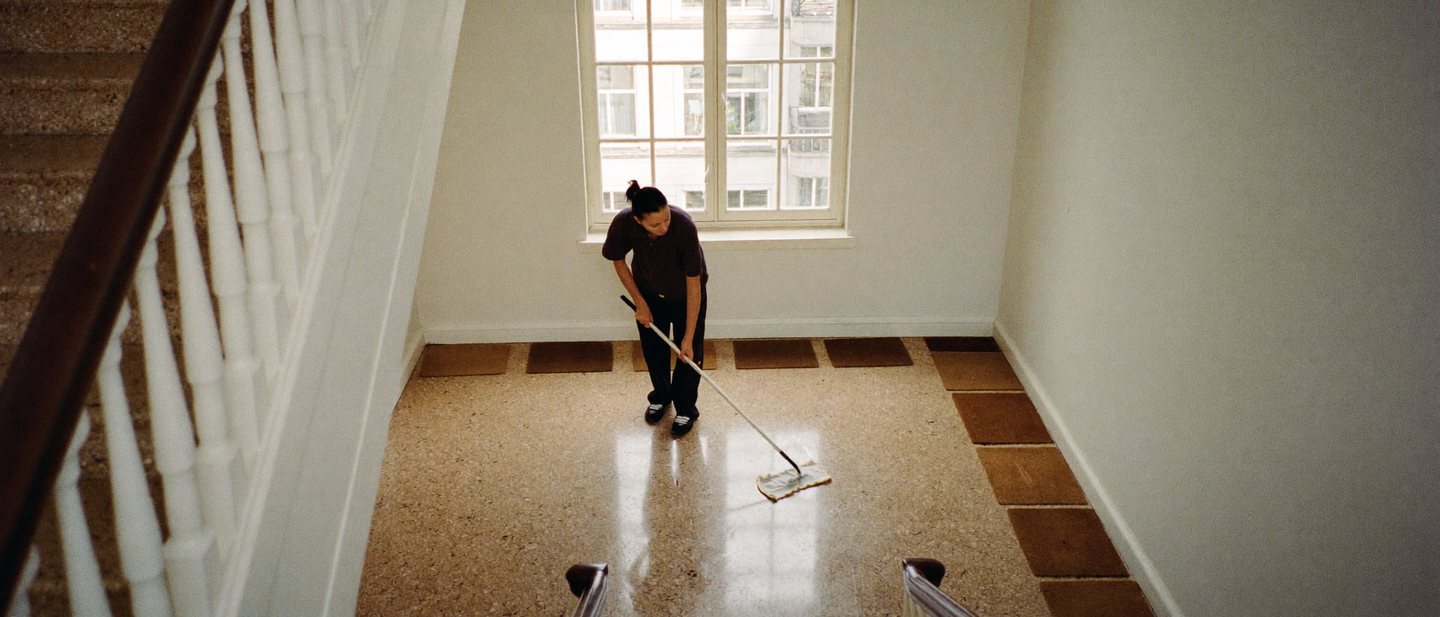 Seen from above, a cleaner mops the shining terrazzo floor of a bright stairwell in a Dutch apartment building, beneath a tall window.
