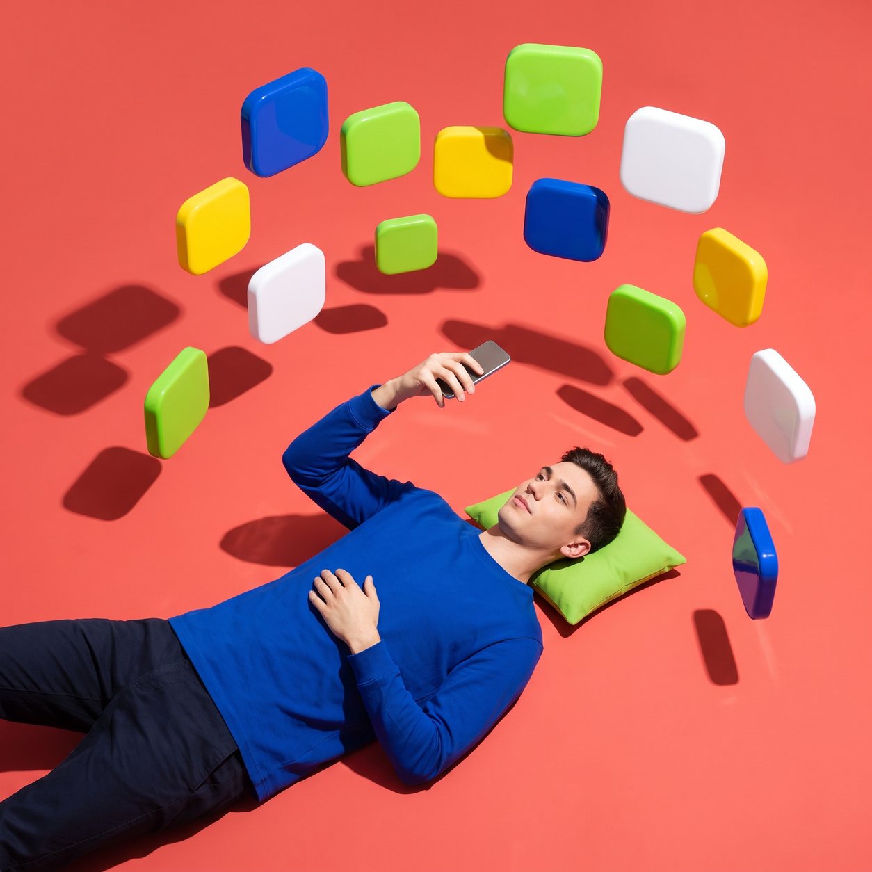 Seen from above, a man lies on a coral-red floor holding a phone, while glossy rounded tiles in cobalt, lime, yellow and white float around him like app icons.