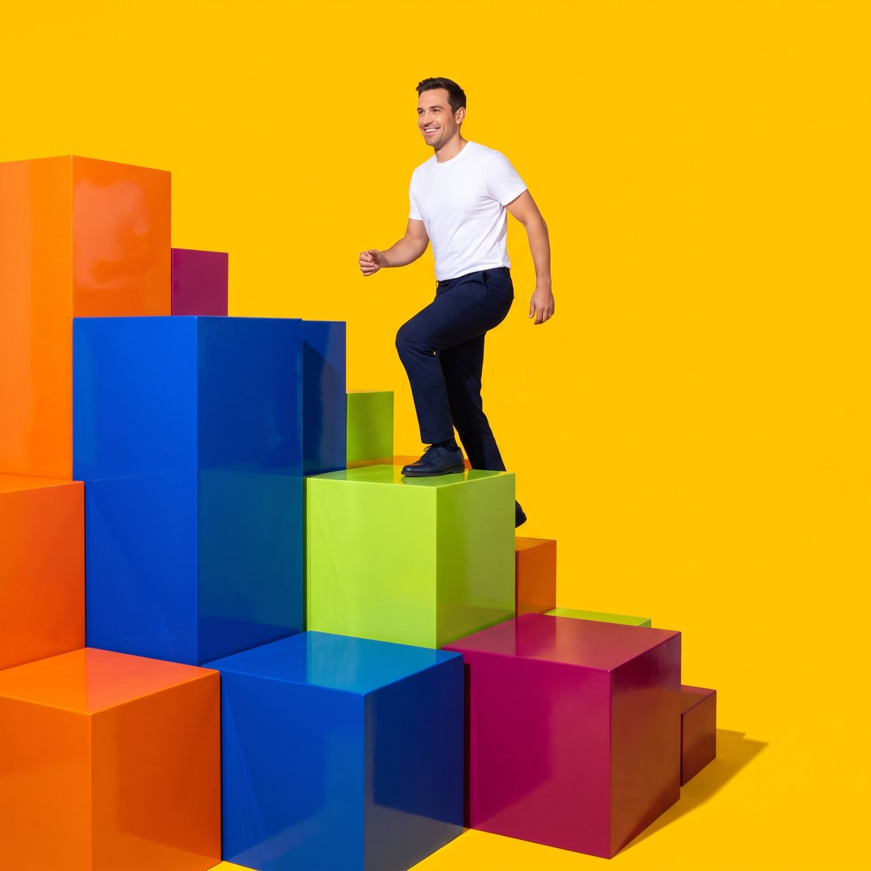 A man in a white t-shirt strides up a staircase of large glossy colour-block cubes, against a sunny-yellow backdrop.